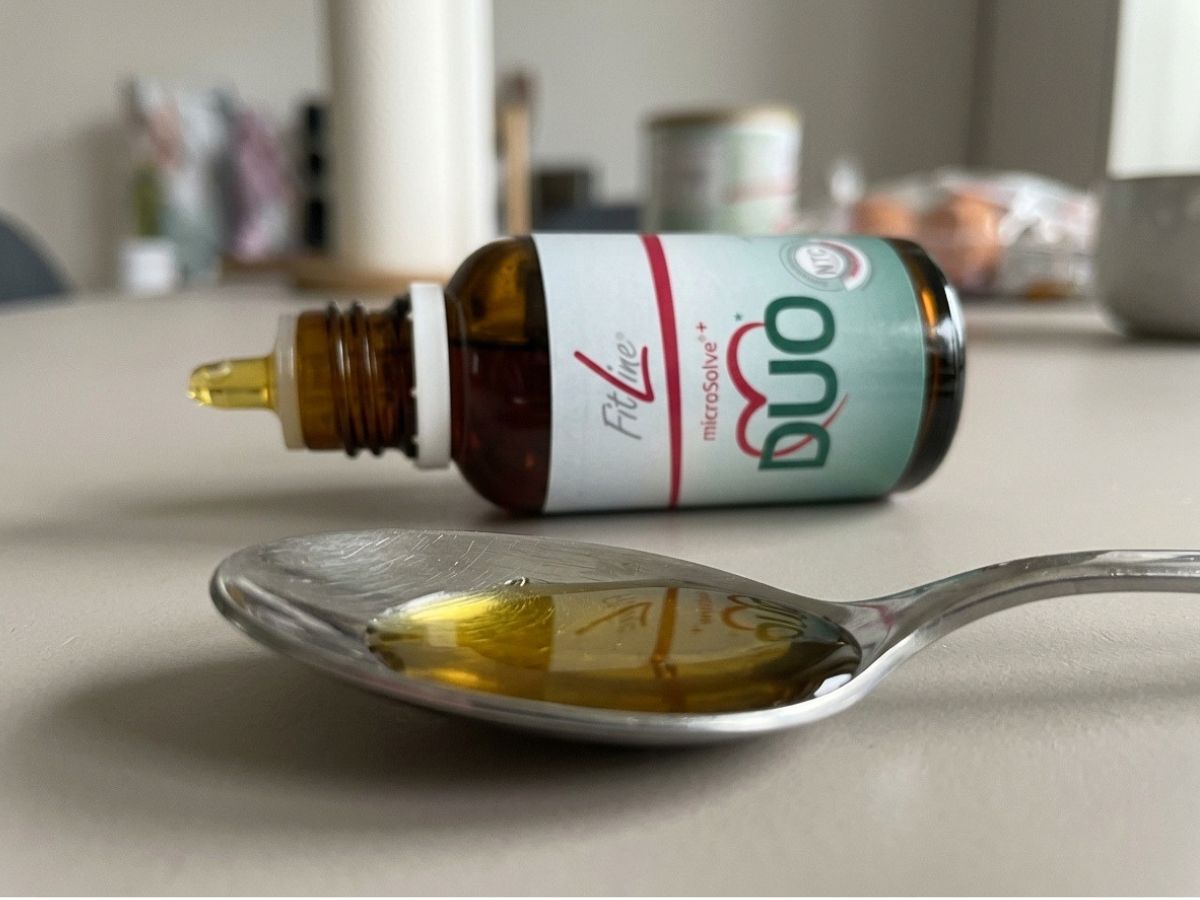 Heart Duo FitLine: gocce oleose giallo-arancio su cucchiaio di acciaio, flacone aperto in secondo piano sul tavolo